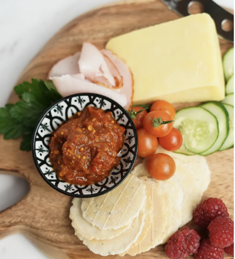 Jar of 'Mumma Made' relish with tomatoes and a glass in the background on a white surface.