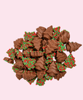A pile of chocolate-covered candy pieces shaped like Christmas trees, decorated with red and green sprinkles, set against a solid pale pink background.