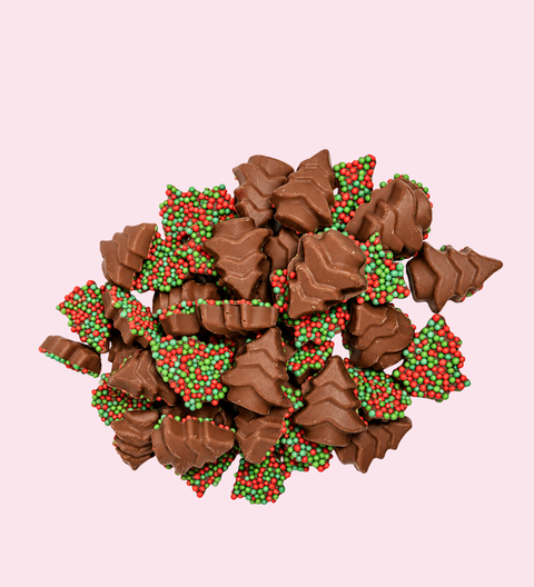 A pile of chocolate-covered candy pieces shaped like Christmas trees, decorated with red and green sprinkles, set against a solid pale pink background.